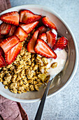 Bowl of muesli, yoghurt and ripe organic strawberries with spoon on a grey surface next to a pink napkin against a blurred background from above