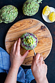 Women's hands halve an artichoke on the stem with a knife on a wooden chopping board