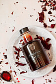 A labelled bottle of hibiscus syrup on a plate with dried hibiscus flowers scattered around.