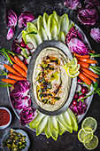 Creamy hummus on oval plate with garnishes on platter with colorful vegetable crudite on dark background