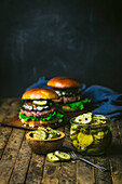 Jar of homemade pickle slices, in a wooden bowl with 2 burgers in the background. Wooden table and chalk in the background