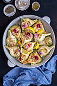 Quarters of artichokes and lemon slices in a round baking pan on a dark background.