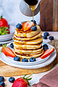 Maple syrup being poured over a stack of pancakes garnished with blueberries and strawberries.