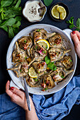 A woman holds a round baking dish filled with fried artichokes, garnished with mint leaves and lemon slices
