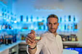 Weichzeichner eines männlichen Barkeepers, der ein Glas mit süßem Schnaps anbietet, während er am Tresen einer modernen Bar steht, vor einem unscharfen blauen Regal mit Flaschen