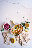 Chickpea hummus served with olive oil, pickled red onions, lemons and flatbread on a white marble table. Female hand dipping flatbread