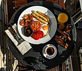 Full fry up English breakfast with fried eggs, sausages, bacon, beans, toasts and coffee served on round table on sunny summer morning
