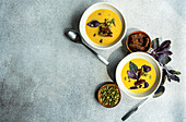 Top view of bowls with pumpkin cream soup with basil herb, rye bread and seeds on blurred grey background with leaves