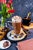 Heiße Schokolade mit Schlagsahne in einem Latte-Glas, Blaubeeren und Schokoladenstückchen drum herum, dahinter Blumen