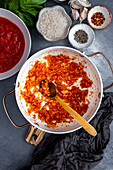 Diced onion beind cooked with tomato paste in a white pot, a wooden spoon inside it, chopped tomatoes, spices, rice, basil and garlic cloves on the side.