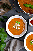 Roasted tomato soup garnished with feta cheese and basil in two white bowls and the soup pot on the side.