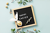 Stylish Easter flat lay with letterbox, egg in Easter bunny napkin, golden eggs, feathers and spring flowers on blue background. Minimalist modern Easter background. Top view flat lay