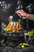 Männliche Hand, die Glasur auf einen Limettenkuchen auf einem gläsernen Kuchenständer mit Gewürzen, Limetten und Cocktailgläsern tropft
