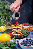 Maple syrup being poured over a stack of pancakes topped with blueberries and raspberries.