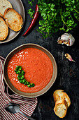 Gazpacho garnished with parsley in a plate with wavy edges. There is red pepper, tomato, garlic, croutons and a spoon on the table. Top view.