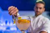 Focused male bartender garnishing a slice of orange on the rim of a cocktail glass with tongs while looking down and standing at the bar in front of blurred blue lights and bottles