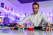 Male bartender serving refreshing whisky and negroni cocktails in glasses while standing behind the counter looking at blurred shelves of bottles