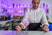 Unidentified male bartender with orange slice in hand looking at the counter with a purple cocktail in the glass while standing in front of a blurred shelf