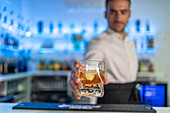 Male bartender standing behind the counter serving a clear glass of cold, refreshing whiskey with ice while looking down and working at the bar against a blurred purple background