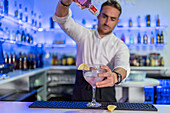 Focused young male bartender pouring an alcoholic drink from a bottle into a cocktail glass with an orange slice on the counter while looking down at the blurred bar