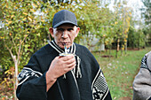 Ernster älterer Mann in gestrickter Mapuche-Kleidung und Mütze schaut in die Kamera, während er Yerba Mate mit einem Strohhalm im grünen Park von Temuco trinkt