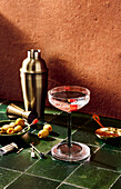 A cocktail glass and cocktail shaker on a bar with olives