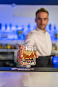 Male bartender standing behind the counter serving a clear glass of cold, refreshing whiskey with ice while looking down and working at the bar against a blurred purple background
