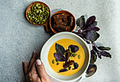 Bowl of cream of pumpkin soup with basil herb, rye bread and seeds on a grey background, viewed from above