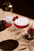 Clover club cocktail in a tall coupe glass garnished with three raspberries next to a shaker in hard light.