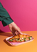 Cropped, unrecognisable person eating a delicious croissant with ham, cheese and rocket on a plate against a colourful background