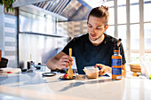 Concentrated young male sitting at table with sushi in plate and looking down while seasoning tuna fish slice with wooden spoon and preparing to serve