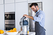 Side view of smiling adult male chef in apron looking down while standing at kitchen table with juice squeezer and fresh cut orange halves in bowl during healthy breakfast process in daylight