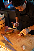 Ein Sushi-Koch stellt ein Stück Nigiri-Sushi an einer warm beleuchteten Restauranttheke sorgfältig zusammen und zeigt dabei kulinarische Präzision