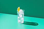 A minimalist shot of a clear glass filled with gin tonic and a slice of lime, casting a shadow on a green background.