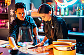 Young focused female chef in black uniform and gloves cutting meat with knife on chopping board while working with male coworker at illuminated sushi bar