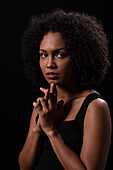 Serious young Hispanic female with curly hair looking at camera and clasping hands against black background
