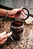 Nahaufnahme einer anonymen Hand, die ein mit fein gemahlenem Kaffee gefülltes Glas öffnet, wobei die Textur des Kaffees und das Armband am Handgelenk vor einer Granitarbeitsplatte hervorgehoben werden