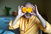Cheerful senior man in casual clothes smiling and covering eyes with halved orange slices while sitting n kitchen