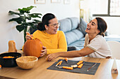 Cheerful young Hispanic female friends looking away while sitting at table with Halloween kit bowls and carving eye with sharp tool on orange pumpkin