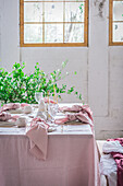 Served table with tablecloth and dishware placed near glasses for banquet in light room decorated by green potted plant