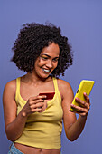 Cheerful Hispanic female with Afro hairstyle in yellow top holding credit card and browsing smartphone while standing on blue background
