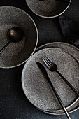 An artistic shot of a modern tableware set, including plates and utensils, arranged on a dark, textured surface.