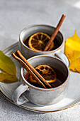 Zwei Tassen mit aromatischem Gewürztee mit Zimtstangen, Anis und getrockneten Orangenscheiben, umgeben von leuchtend gelben Herbstblättern auf strukturiertem grauem Hintergrund
