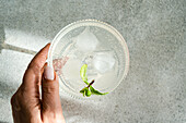 Close-up of a transparent drink with ice, lime slice, and mint in a glass cup, held by a person's hand over a grey surface