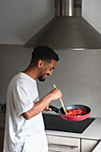 Side view of positive young ethnic African American male in casual clothes preparing delicious dish while frying tomatoes on pan and mixing ingredients with wooden spatula during cooking process in kitchen