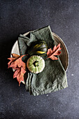 Draufsicht auf einen herbstlich gedeckten Tisch mit Serviette, Blättern und Kürbissen auf einem Keramikteller vor dunkler Oberfläche