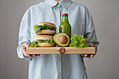 Crop anonymous woman in shirt holding healthy green burgers with lettuce on wooden cutting board while standing over grey background