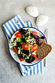 Draufsicht auf einen saisonalen Gemüsesalat mit Bio-Gurke, Tomate, Zwiebel und roten Basilikumblättern, serviert auf einem Teller mit Besteck auf einem Holztablett neben einer Serviette vor grauem Hintergrund