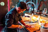 Side view of smiling male chef wearing gloves and black uniform slicing fish with knife on illuminated wooden counter at modern sushi bar