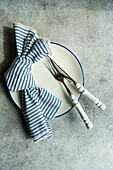 Draufsicht auf einen minimalistisch-rustikal gedeckten Tisch mit weißem Teller, Besteck und gestreifter Serviette auf grauem Untergrund